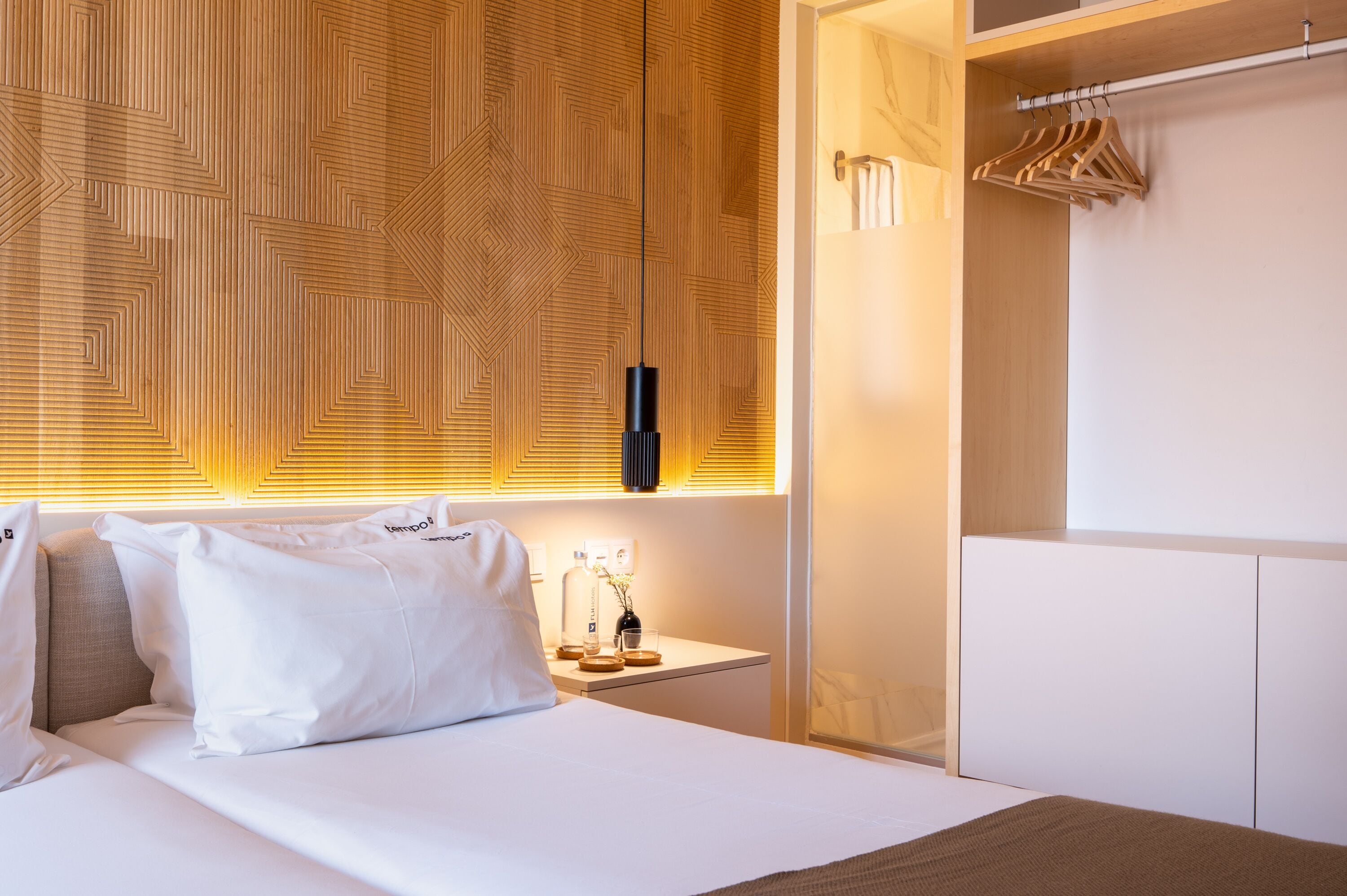 Quarto de hotel com cama, mesa de cabeceira com luminária, espelho e guarda-roupa com cabides de madeira.