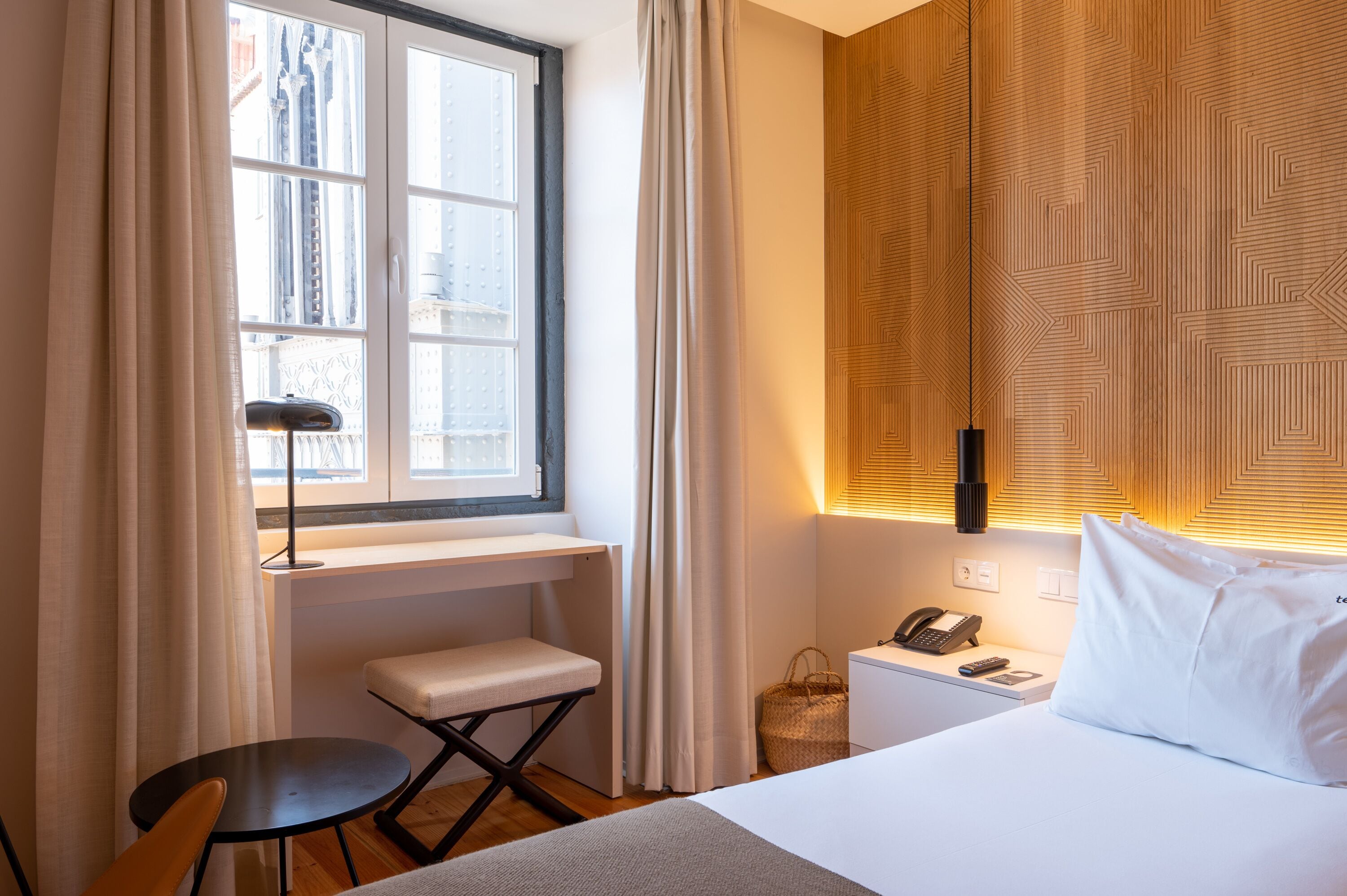 Une chambre d’hôtel comprenant un lit, un bureau, une chaise, une petite table, une fenêtre avec des rideaux, un téléphone fixé au mur et une lampe de chevet.