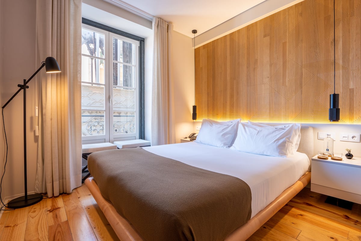 Een moderne hotelkamer met een groot bed, nachtkastje, lamp en uitzicht door een raam.
