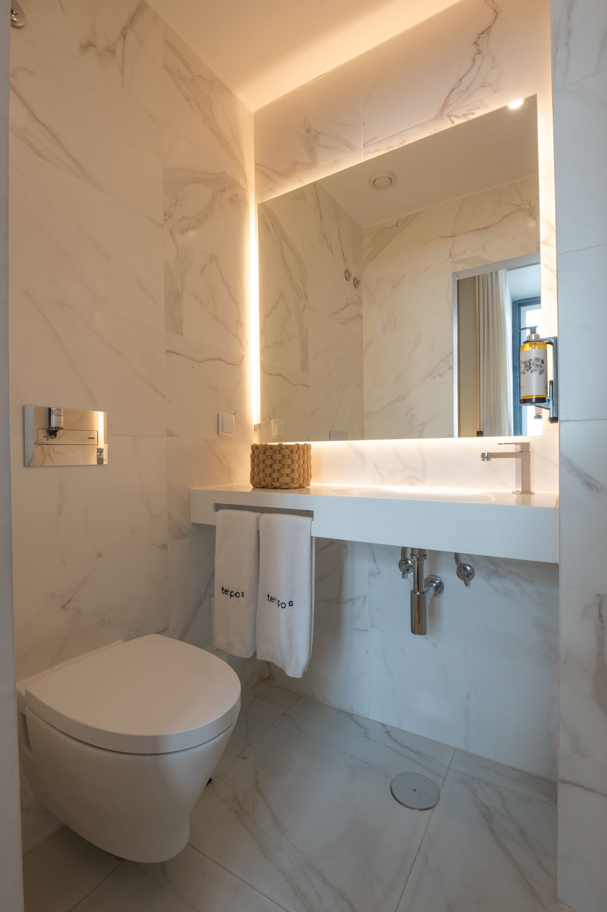 A modern bathroom with a marble floor, a white toilet, a sink with a mirror above it, and a towel rack with two towels.