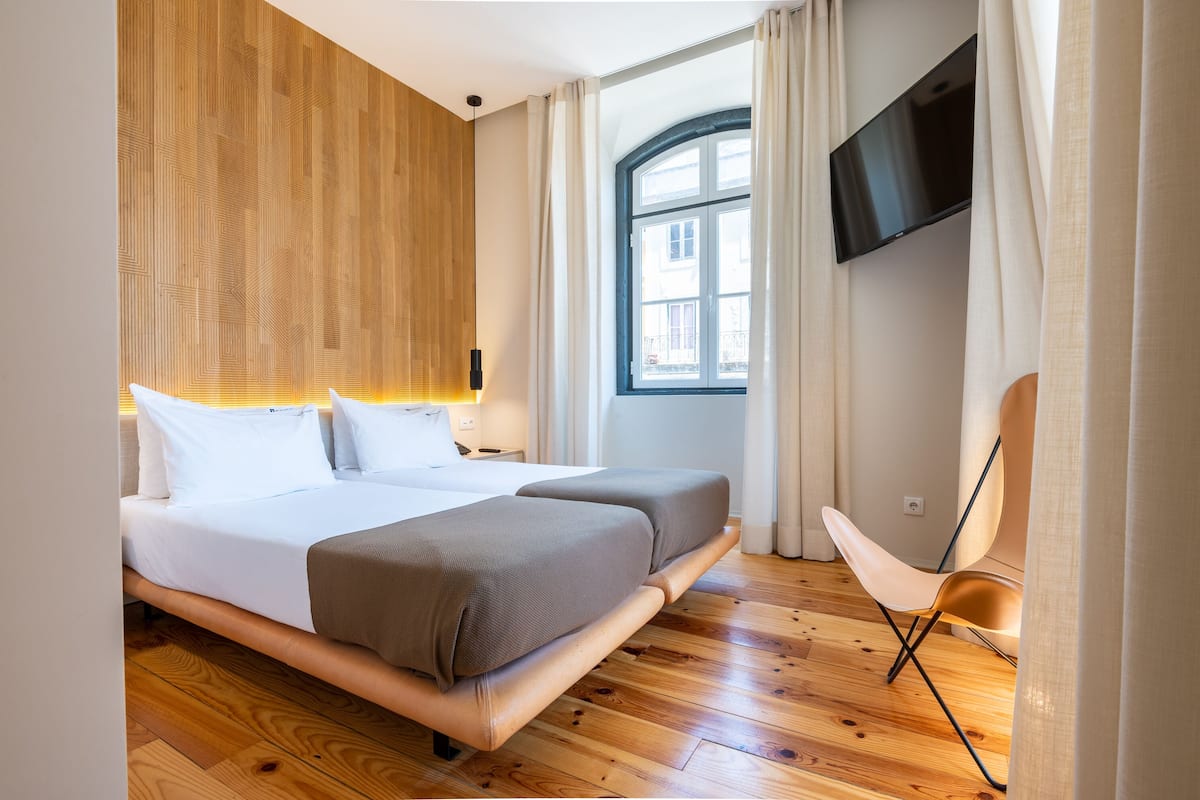 Een moderne hotelkamer met een houten vloer, een groot bed met twee kussens, een nachtkastje met een lamp, een aan de muur bevestigde televisie en een raam met gordijnen.