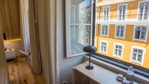 Een kamer met een houten vloer, een bureau met een lamp, een stoel en een raam met uitzicht op een geel gebouw.