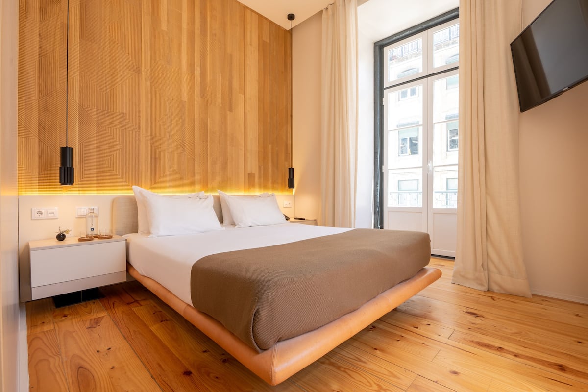 Een moderne slaapkamer met een houten hoofdeinde, een groot bed, een nachtkastje, een flatscreen-tv en een raam met gordijnen.