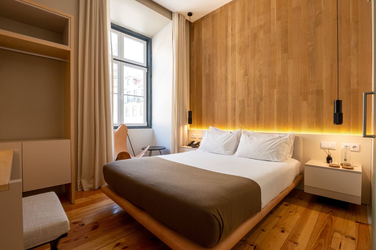Een moderne hotelkamer met een groot bed, houten vloer, een inbouwkast, nachtkastje met lamp en een raam met gordijnen.
