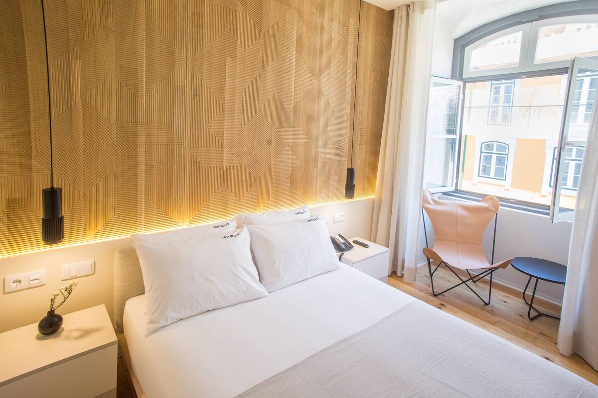 Een moderne hotelkamer met een groot bed, een nachtkastje, een stoel en een raam met uitzicht op gebouwen.