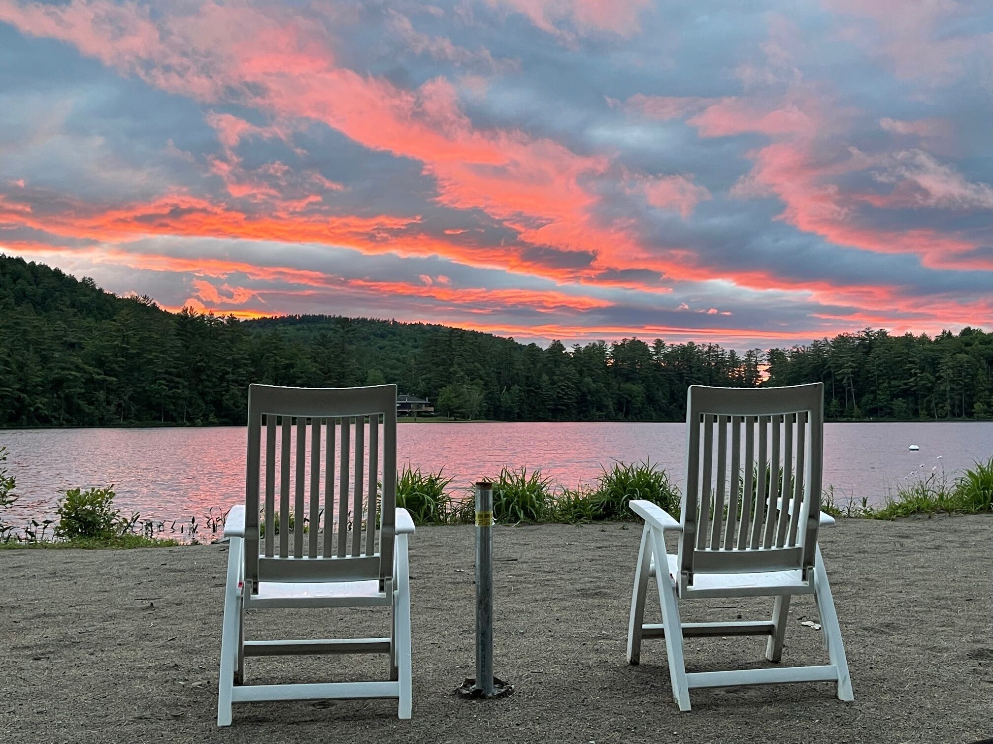 Deux chaises Adirondack blanches, placées face à un lac, avec une colline boisée en arrière-plan et un ciel au coucher du soleil.
