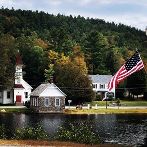 A small stone building with a red door, a church with a red steeple, and an American flag on a flagpole.
