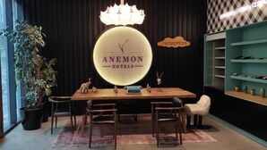 Ein Empfangsbereich mit einem Holztisch, Stühlen, einem großen beleuchteten Schild mit der Aufschrift „ANEMON HOTELS“ und einer Rezeption.