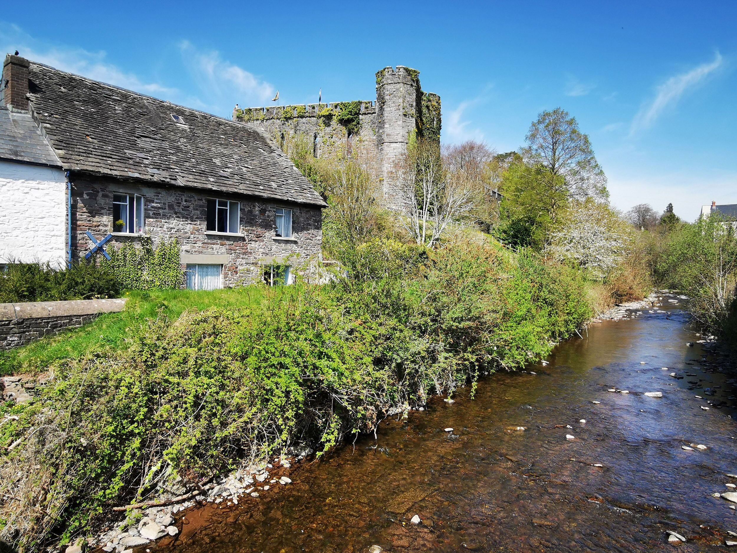 A stone house with a river in front, surrounded by greenery and a castle in the background.