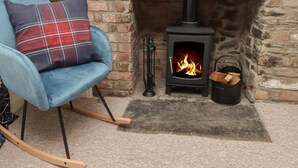 A cozy room with a brick fireplace, a blue armchair with a plaid cushion, and a black wood stove. - Kooky Cottage (Brecon)