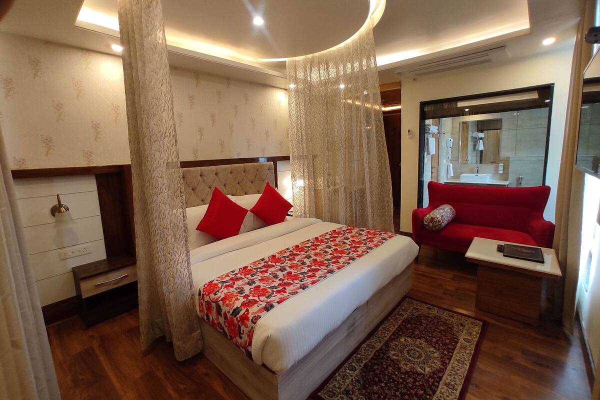 A modern hotel room with a large bed, a red sofa, a small table, and a bathroom visible through a glass door.