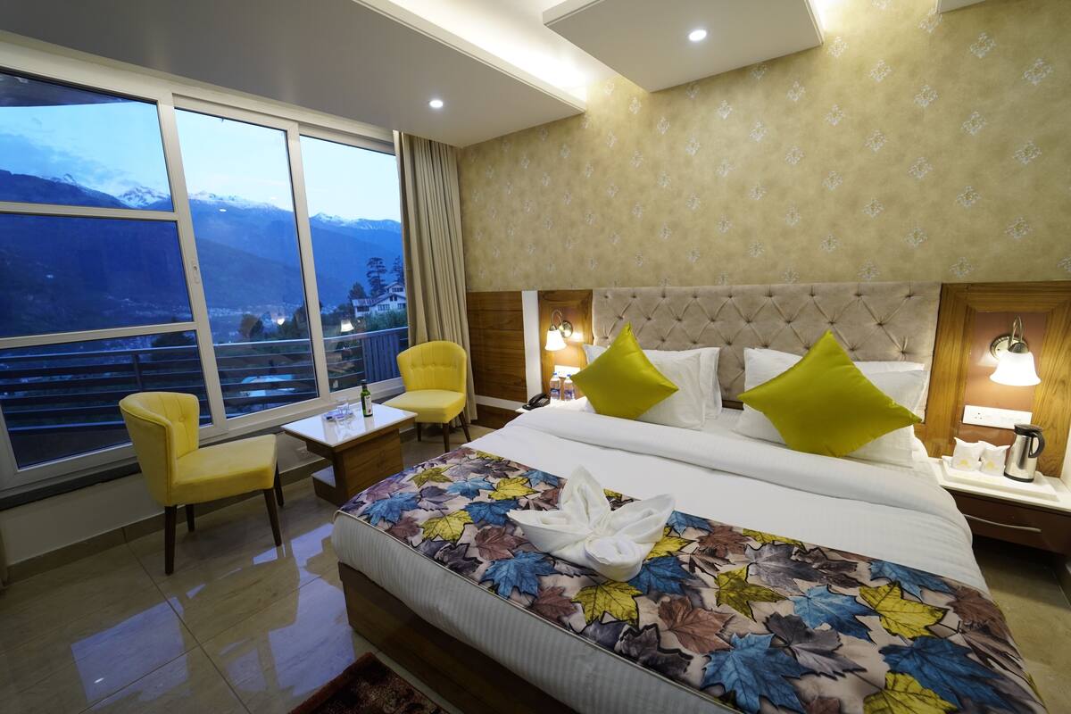 A hotel room with a bed, a chair, a small table, and a view of mountains through large windows.