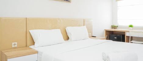 A neatly made bed with white linens and a rolled white towel, a wooden nightstand with a white drawer, and a small desk with a plant.