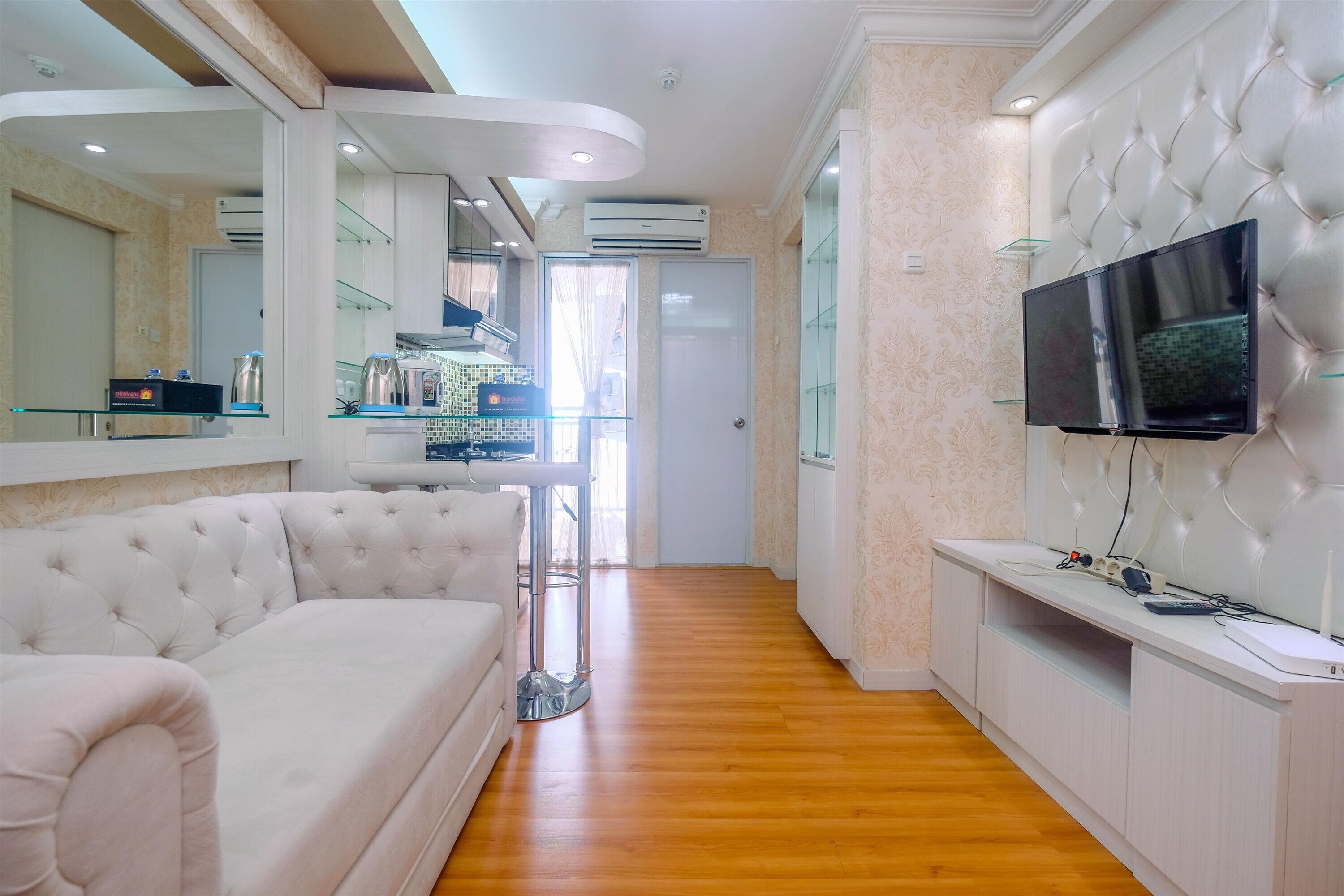 A modern living room with a white sofa, a glass coffee table, a flat-screen TV mounted on a white wall, and a wooden floor.