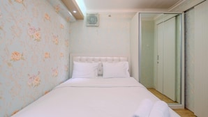 A neatly made bed with white linens and pillows, a tufted headboard, floral wallpaper, and a sliding glass door leading to a walk-in closet. - Cozy Stay 2BR Bassura City Apartment (Jakarta)