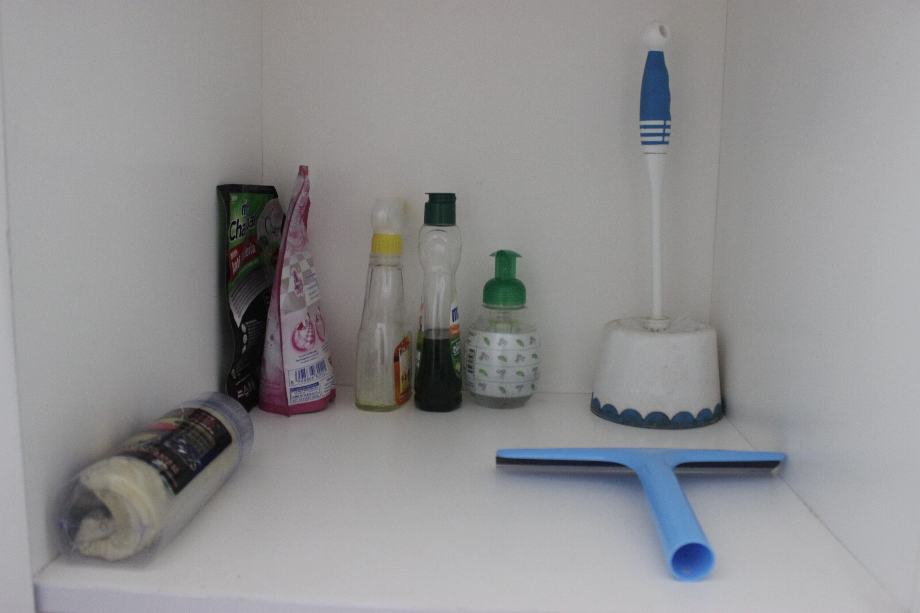 Un estante con productos de limpieza, incluyendo una botella pulverizadora, jabón para platos, jabón de manos, un rollo de papel higiénico y un cepillo de limpieza azul.