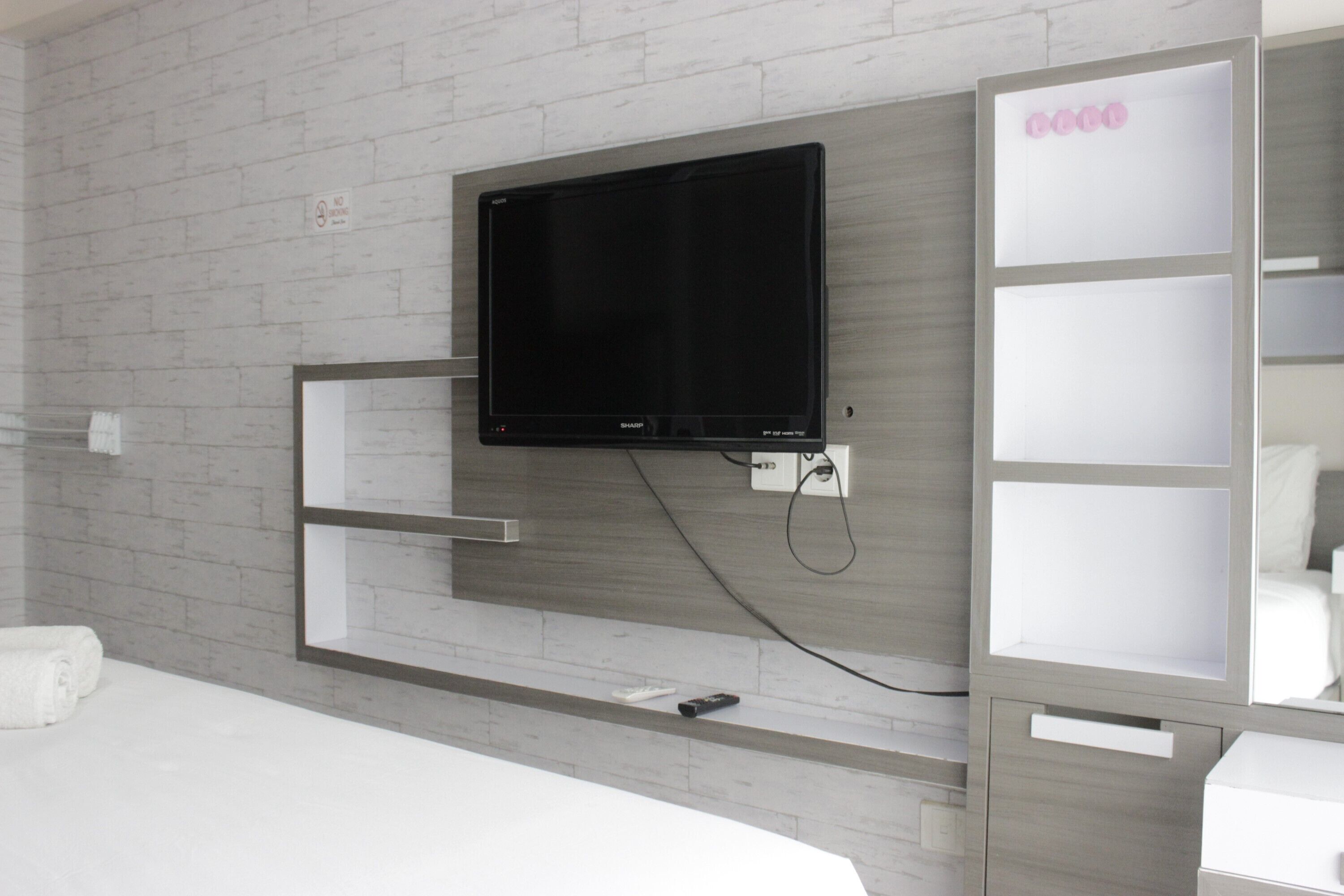 A modern hotel room with a flat-screen TV mounted on a brick-patterned wall, a built-in shelf unit, and a bedside table with a remote control.