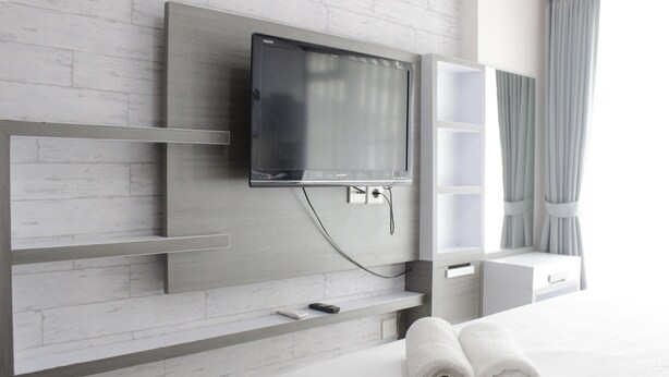 A modern hotel room with a flat-screen TV mounted on a white brick wall, a bed with white linens, and a small shelf with a remote control.