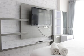 A modern hotel room with a flat-screen TV mounted on a white brick wall, a bed with white linens, and a small shelf with a remote control. - Strategic & Stylish Studio Gateway Pasteur Apartment near Pasteur Exit Toll (Cimahi)