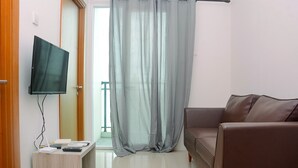 Uma sala de estar com sofá de couro marrom, uma televisão de tela plana montada em suporte de madeira e cortina cinza parcialmente fechada.