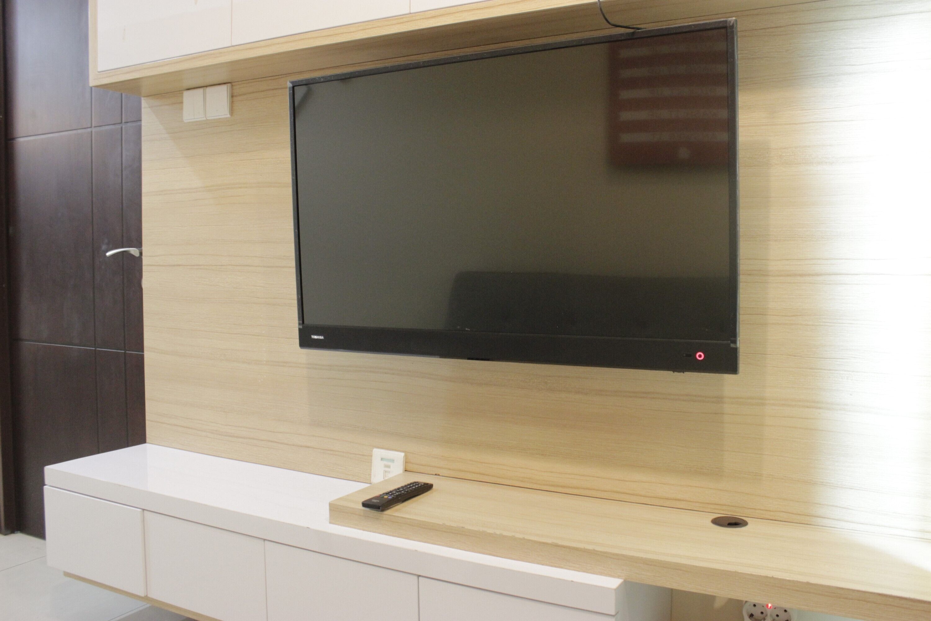 A modern living room with a flat-screen TV mounted on a wooden wall, a white entertainment unit below, and a remote control on the unit.