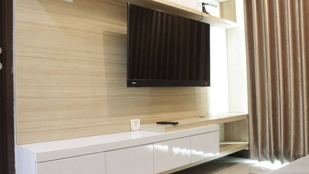 A modern hotel room with a flat-screen TV mounted on a wooden wall, a white console table with a remote control, and a large window with curtains. - Best Location 2BR Tamansari La Grande Apartment (Bandung)