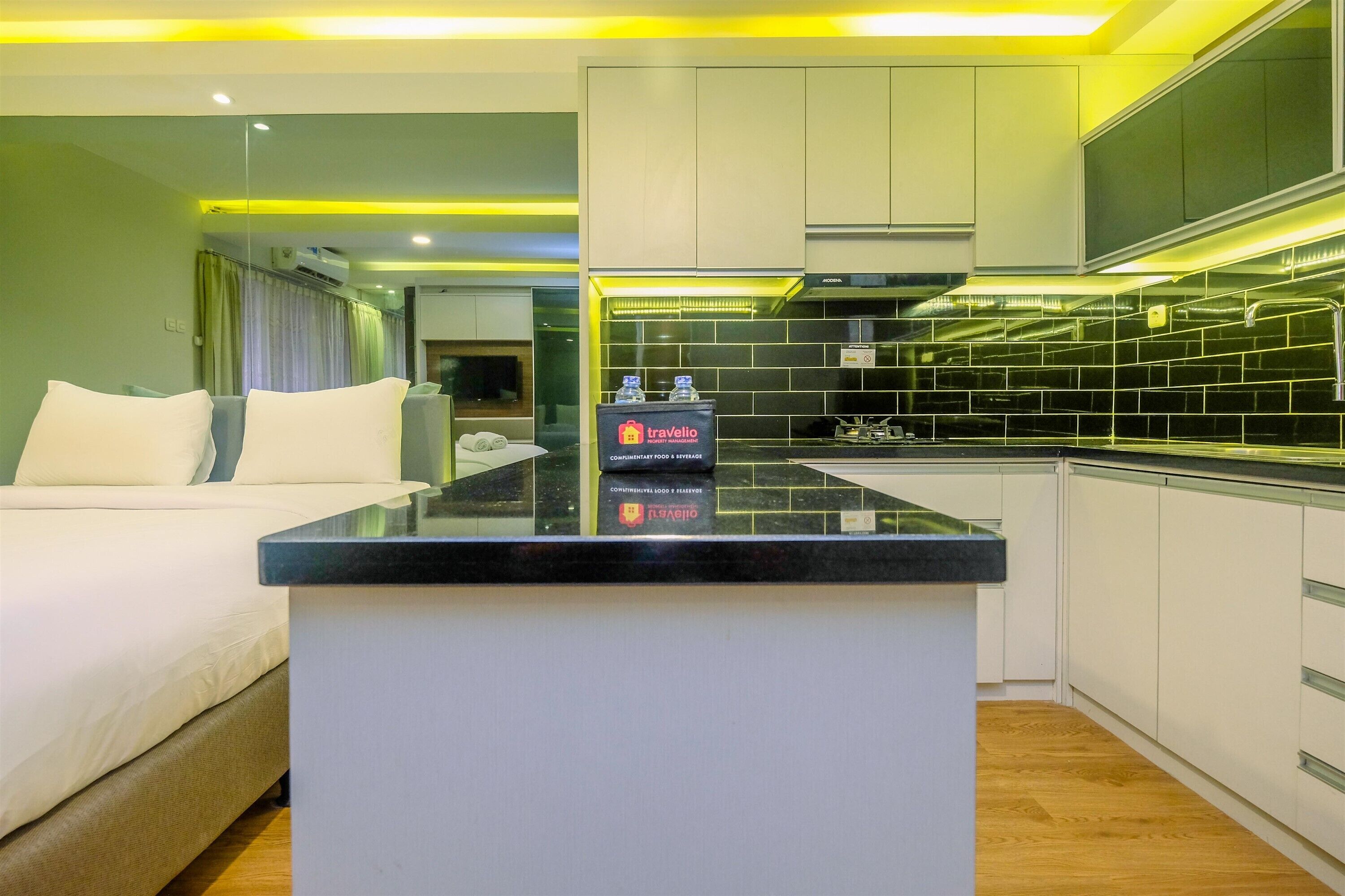 Habitación de hotel moderna con una isla central, cama con ropa blanca y una kitchenette con salpicadero de azulejos negros.