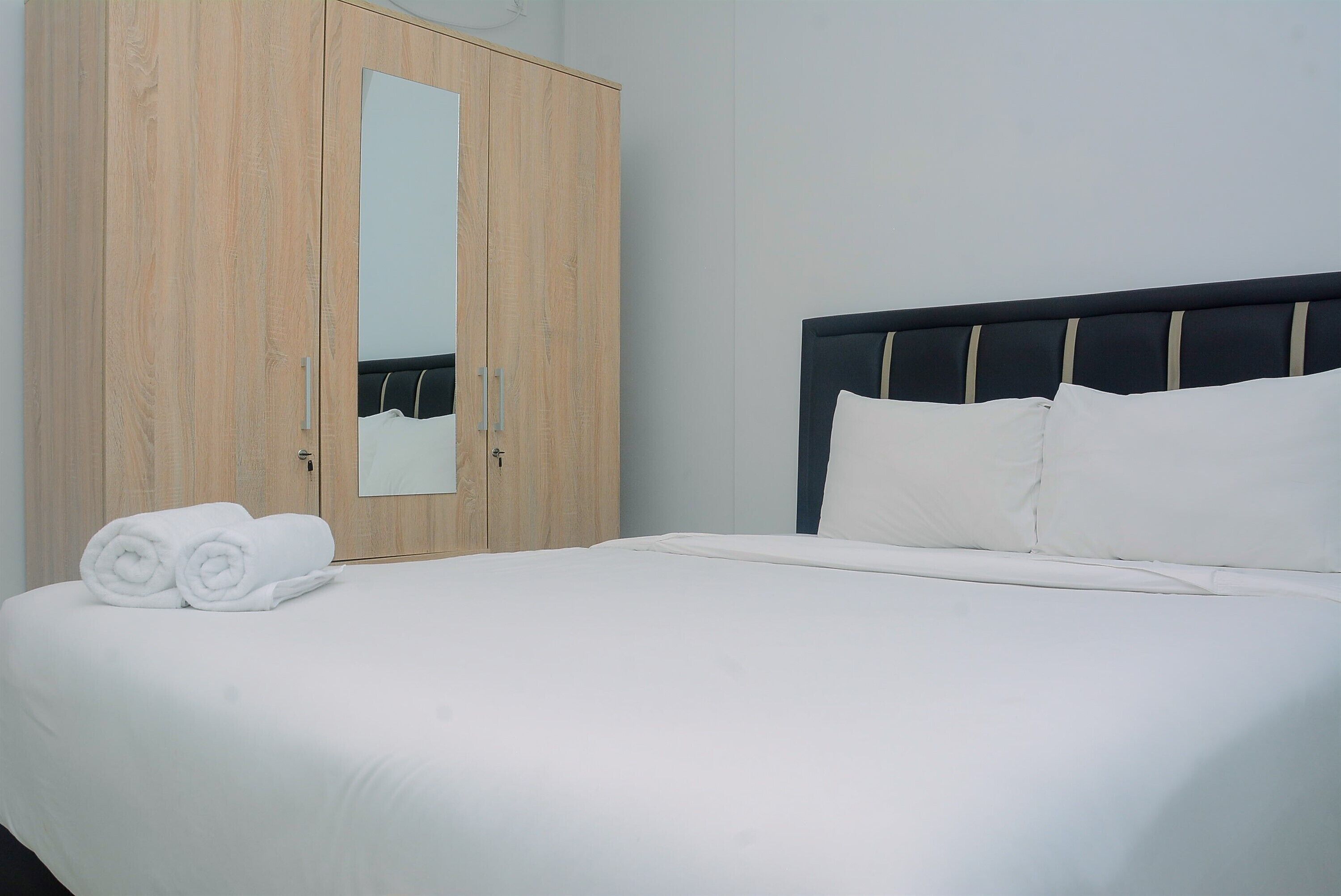 A neatly made bed with white linens and pillows, a wooden wardrobe with a mirror, and a plain wall.