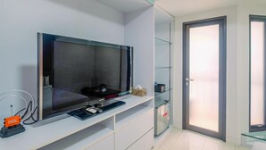 A modern hotel room with a large flat-screen TV on a white console, a glass door with frosted glass panels, and a small shelf with a tissue box and a remote control. - Comfort Studio Tamansari Sudirman Apartment (Jakarta)