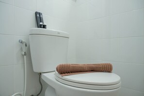 A white toilet with a beige towel on the seat and a bidet attachment on the tank. - Homey Studio at Sentraland Cengkareng Apartment near Airport (Jakarta)