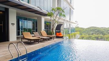 A modern hotel with a rooftop infinity pool, lounge chairs, and a view of lush greenery.
