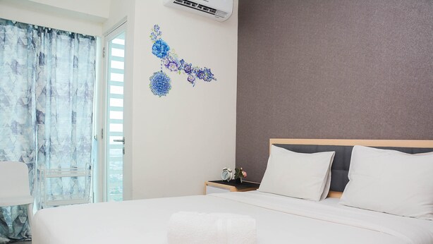 A neatly made bed with a white towel, a bedside table with a clock and a small plant, and a wall with a floral decoration. - Cozy Stay 1BR at Grand Kamala Lagoon Apartment (Bekasi)