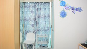 A room with a sliding glass door, a white chair, a white shelf unit, and a wall with floral decorations. - Cozy Stay 1BR at Grand Kamala Lagoon Apartment (Bekasi)