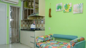 A compact kitchen with a built-in stove, a microwave, and a water dispenser. There is a colorful sofa with a patterned cover and a teal armchair. The walls are painted green, and there are three framed leaf paintings on the wall. - Comfortable 2BR Bassura City Apartment near Bassura Mall (Jakarta)