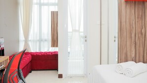 A hotel room with a red sofa, a white bed with rolled towels, a wooden accent wall, and a large window with sheer curtains.