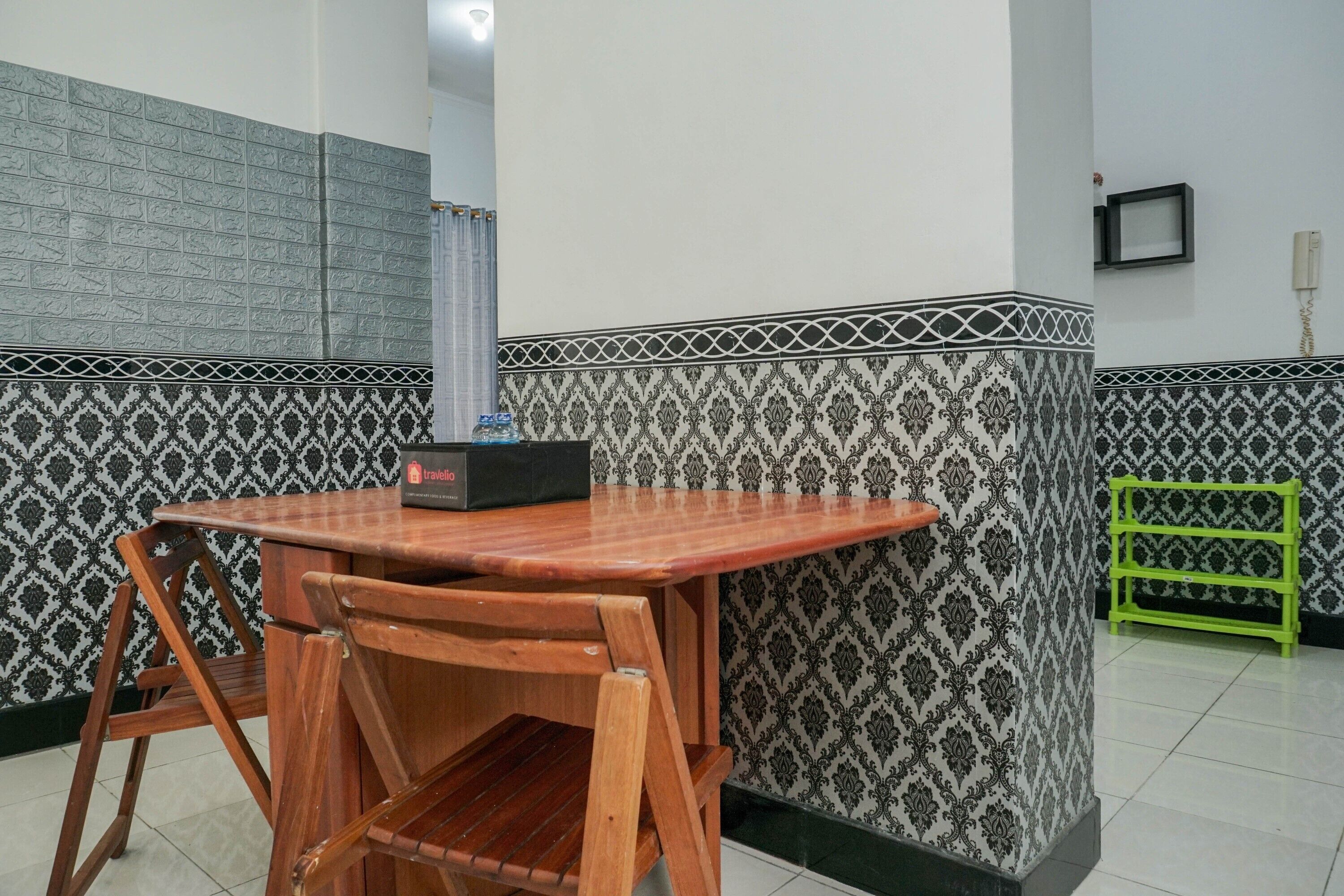 A small dining area with a wooden table and chairs, a black box on the table, patterned wallpaper, and a green rack.