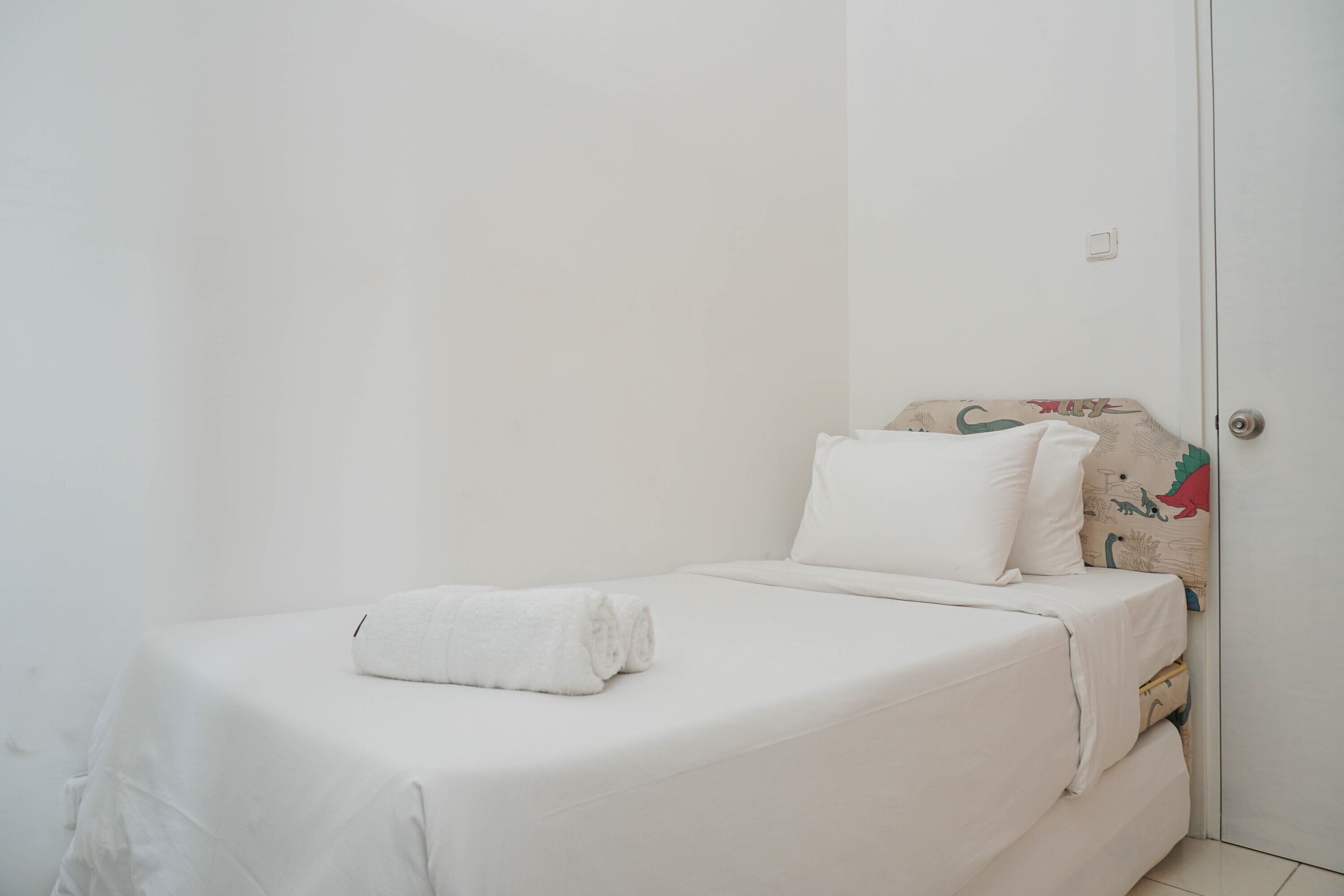 A small, neatly made bed with white linens and a rolled towel on the side, a plain white wall, and a door with a simple handle.