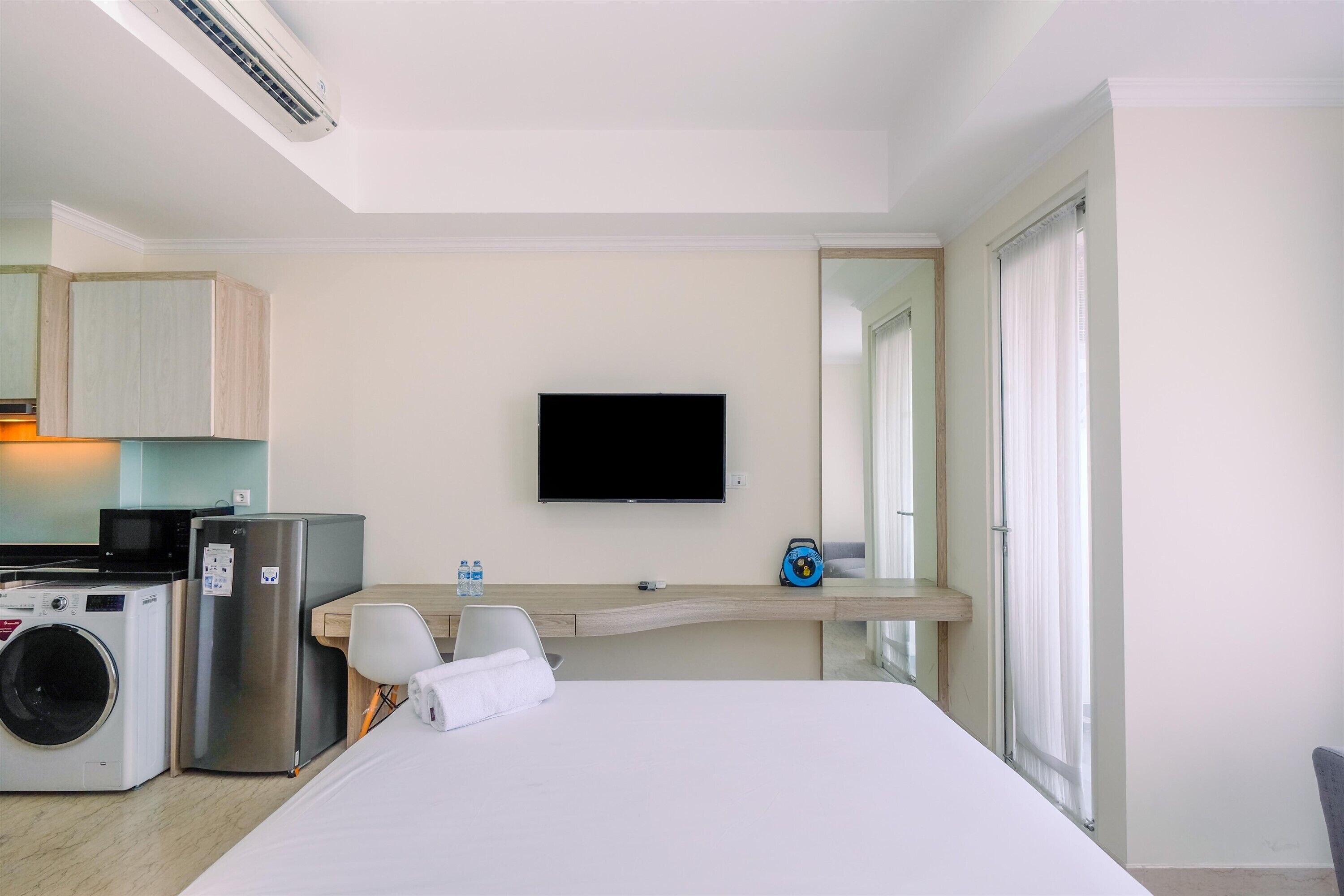 Habitación de hotel con cama, televisor, lavadora, refrigerador, microondas, una pequeña mesa con silla y ventana con cortinas.