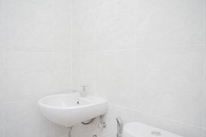 Features a wall-mounted sink with a chrome faucet and a toilet positioned beside it, set against a backdrop of large, white tiles.