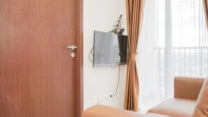 A room with a wooden door, a flat-screen TV mounted on the wall, a window with curtains, and a beige sofa. - Comfy 2BR Apartment at Parkland Avenue (South Tangerang)