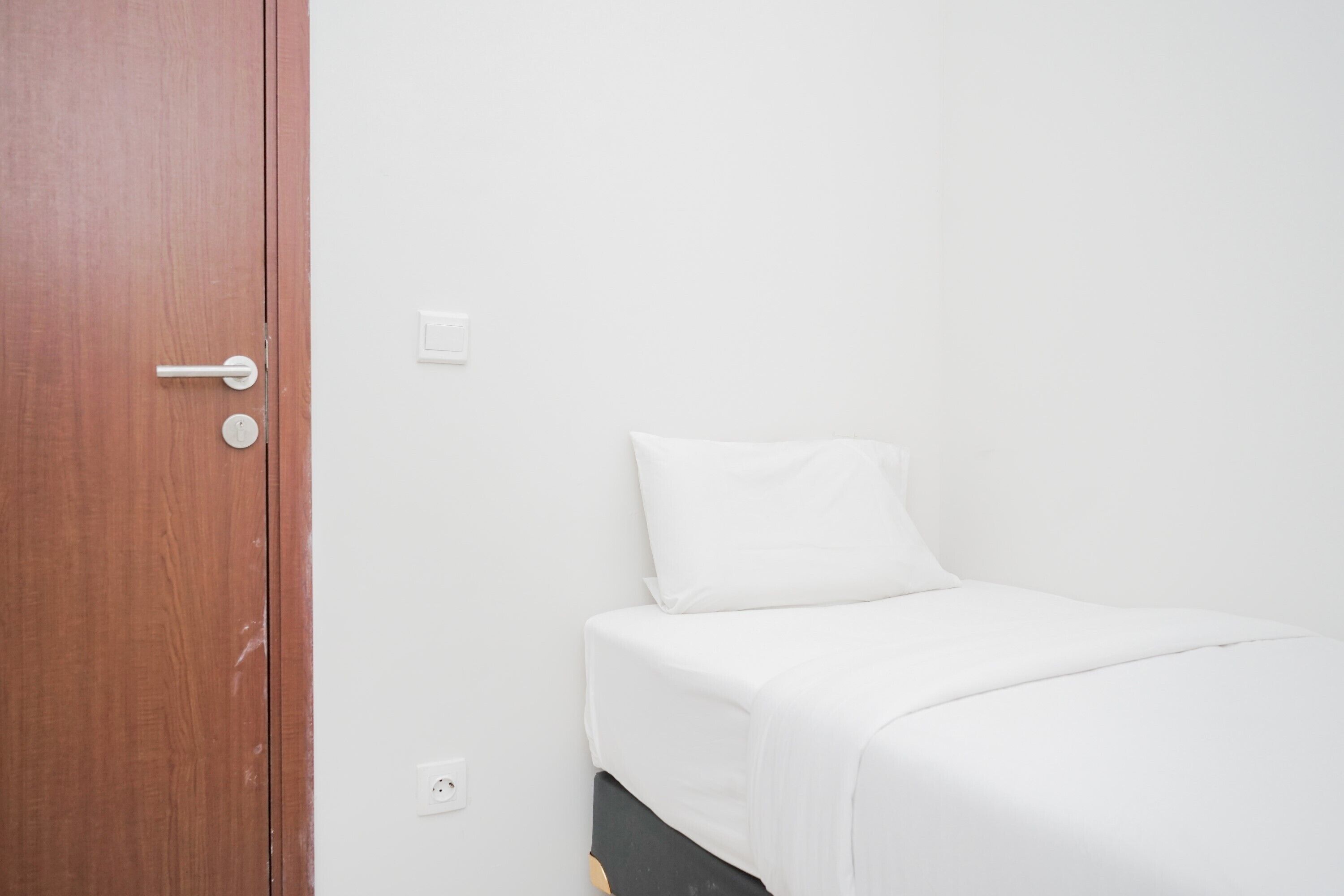 A room with a wooden door, a white wall, and a bed with a white mattress and pillow.