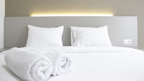 A neatly made bed with white linens and pillows, a rolled white towel, and a light switch on the wall.