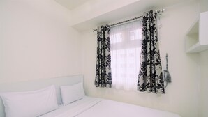 A bedroom with a bed, white bedding, a window with patterned curtains, and a wall-mounted light fixture.
