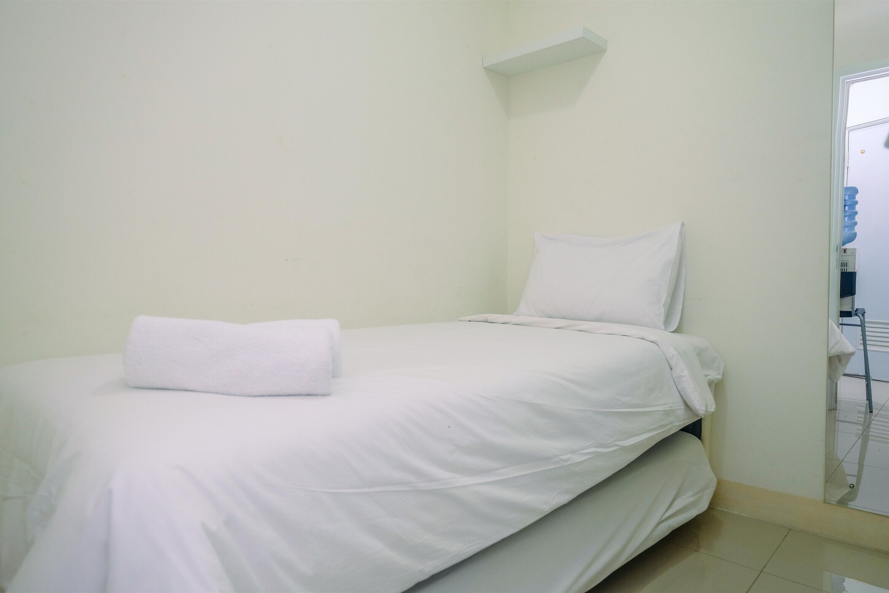 A small, neatly made bed with white linens in a simple room with plain walls and a small shelf above the bed.