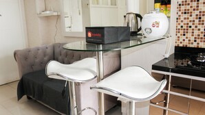 A modern kitchen with a glass-top table, two white bar stools, a grey sofa, and a travelio box on the table. - Comfort 2BR Bassura City Apartment near Shopping Center (Jakarta)