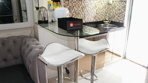 A modern kitchen with a glass countertop, two bar stools, a tufted sofa, and a tiled backsplash. - Comfort 2BR Bassura City Apartment near Shopping Center (Jakarta)