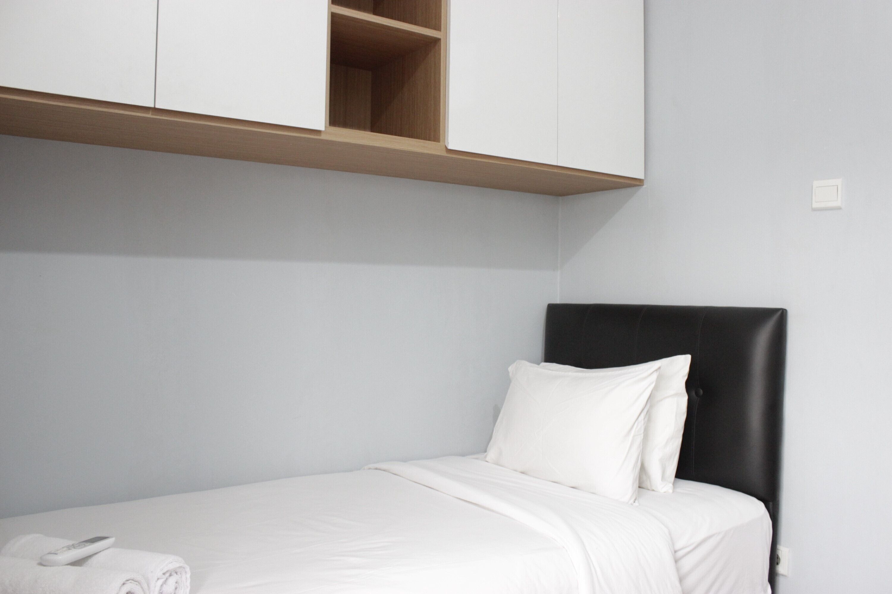 A single bed with a black headboard, white bedding, and a wooden shelf above.