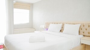Un lit bien fait, avec du linge de lit blanc et des oreillers, une table de chevet avec une lampe, et une fenêtre avec des rideaux de couleur beige.