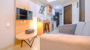 Una sala de estar moderna con un sofá gris, una mesita redonda con un caja, un televisor de pantalla plana montado en la pared y una zona de cocina con microondas y cafetera.