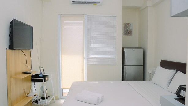 A hotel room with a bed, a TV mounted on a wooden shelf, a refrigerator, and a window with blinds. - Comfort Studio at Green Pramuka Apartment near Shopping Center (Jakarta)